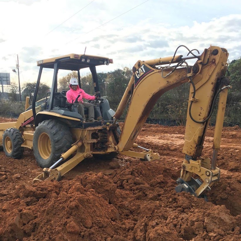 A woman sits on a backhoe in a dirt field.
